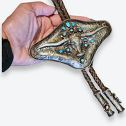 Hand holding a decorative bolo tie with bull design and turquoise stones on a white background